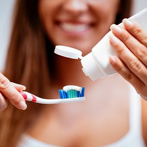 Woman blurry in background applying toothpaste to toothbrush in foreground